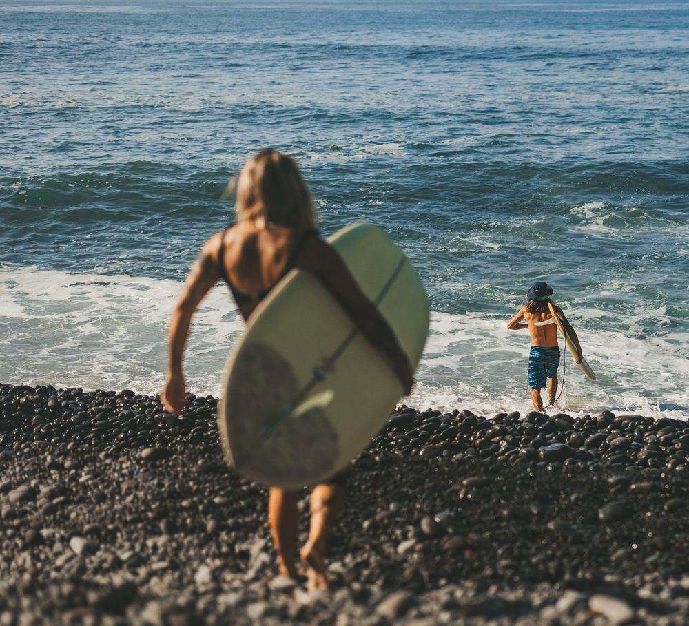 A surfer walking down the cobblestones toward the water with her longboard under her arm