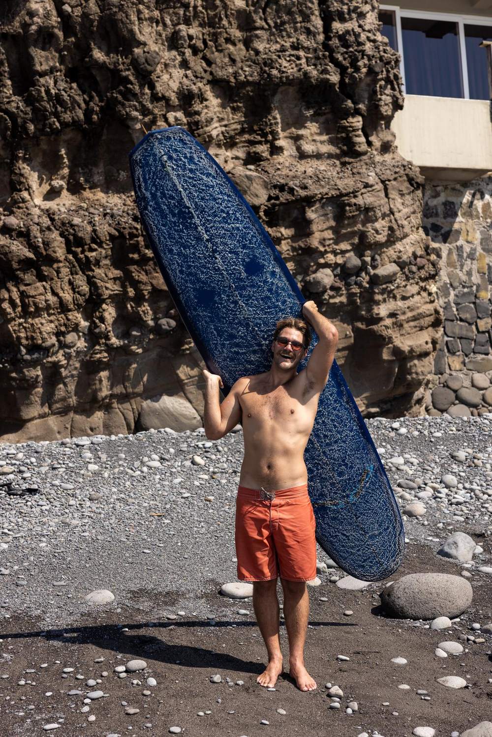 Kevin grinning on a cobblestone beach with his longboard over his shoulder