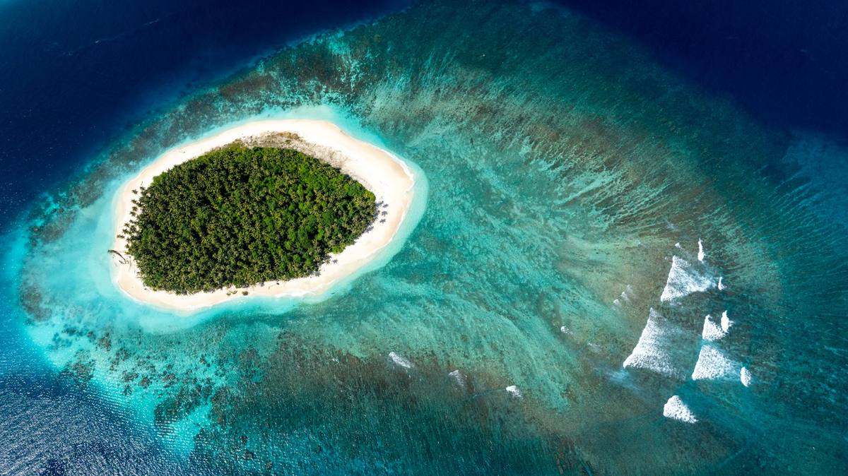 Drone view of a small palm covered island ringed by reef in the Mentawais, sets breaking on the outer reef