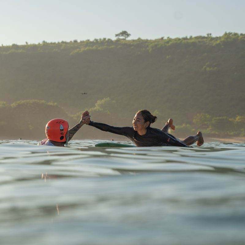 Tommy in the water in a helmet, high fiving a guest on her longboard